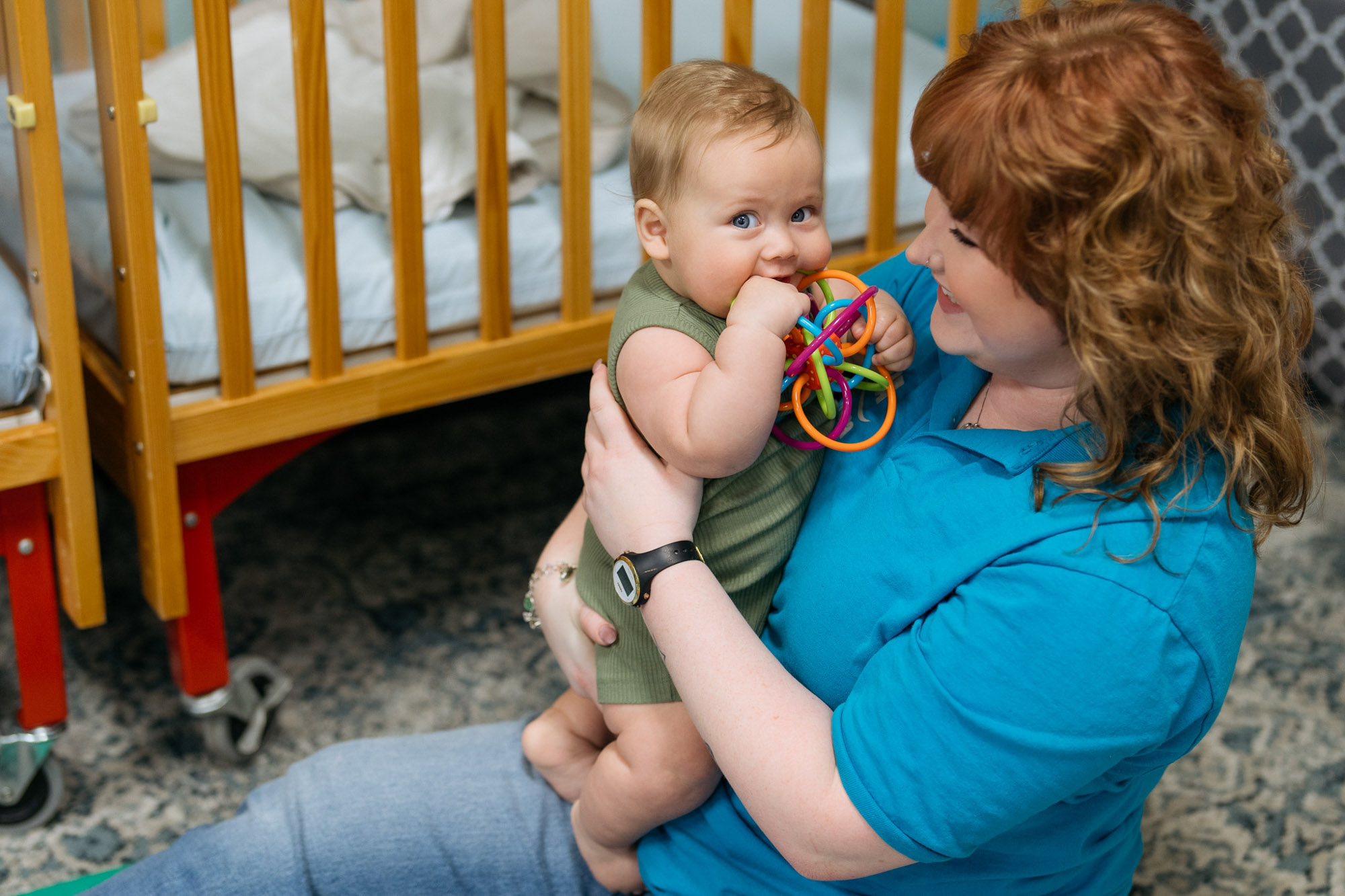 A woman in a blue shirt holds a baby in a green outfit who is chewing on a colorful plastic ring toy near a wooden crib.