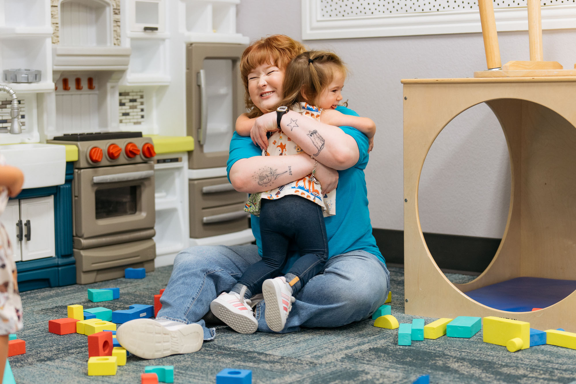 An adult and a child sit on the floor of a playroom, hugging each other amid scattered colorful plastic blocks and play kitchen furniture.