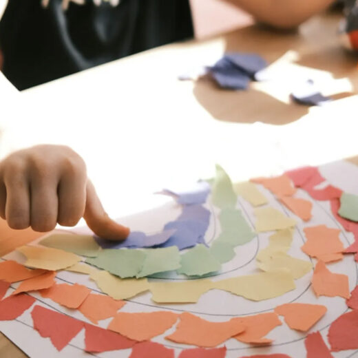 A child’s hand arranges torn colored paper pieces into the shape of a rainbow on a sheet of paper at a table.