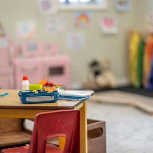 A small table with a red chair holds lunch containers and a milk bottle in a colorful, decorated classroom or playroom.