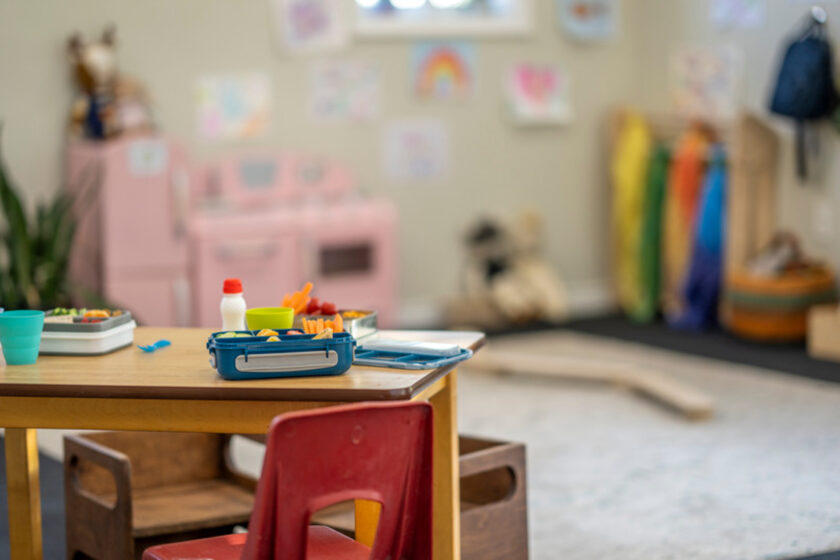 A small table with a red chair holds lunch containers and a milk bottle in a colorful, decorated classroom or playroom.