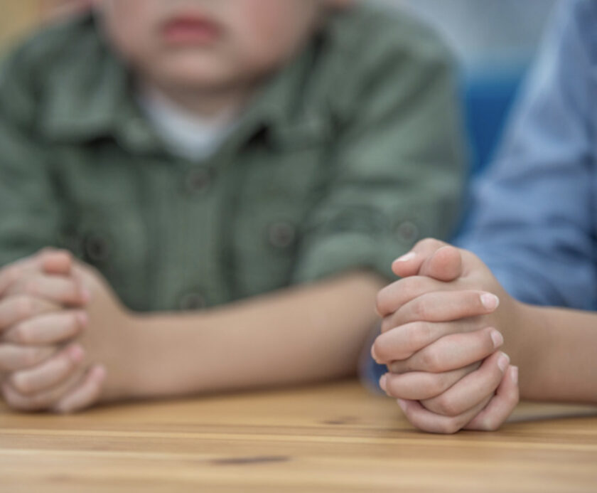 Two children sit at a table with their hands clasped together, shown from the shoulders down. Their faces are not fully visible.