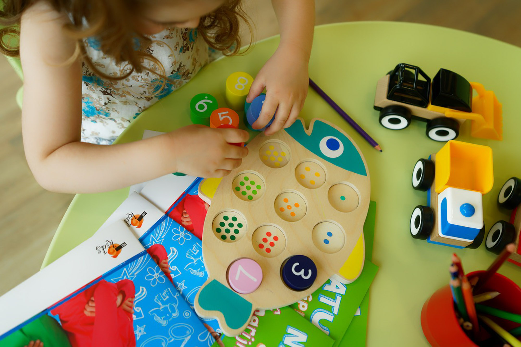 A child plays with a wooden fish-shaped puzzle with numbered pieces on a table, next to toy trucks, pencils, and colorful books.