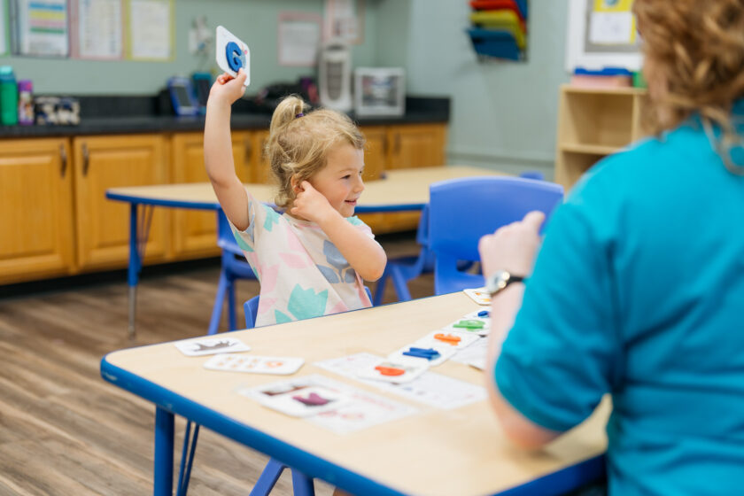 A young girl sits at a classroom table raising a card in her hand, participating in an activity with an adult across from her.
