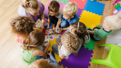 A group of young children sit in a circle on colorful foam mats, looking at alphabet cards with an adult in a classroom or playroom setting.